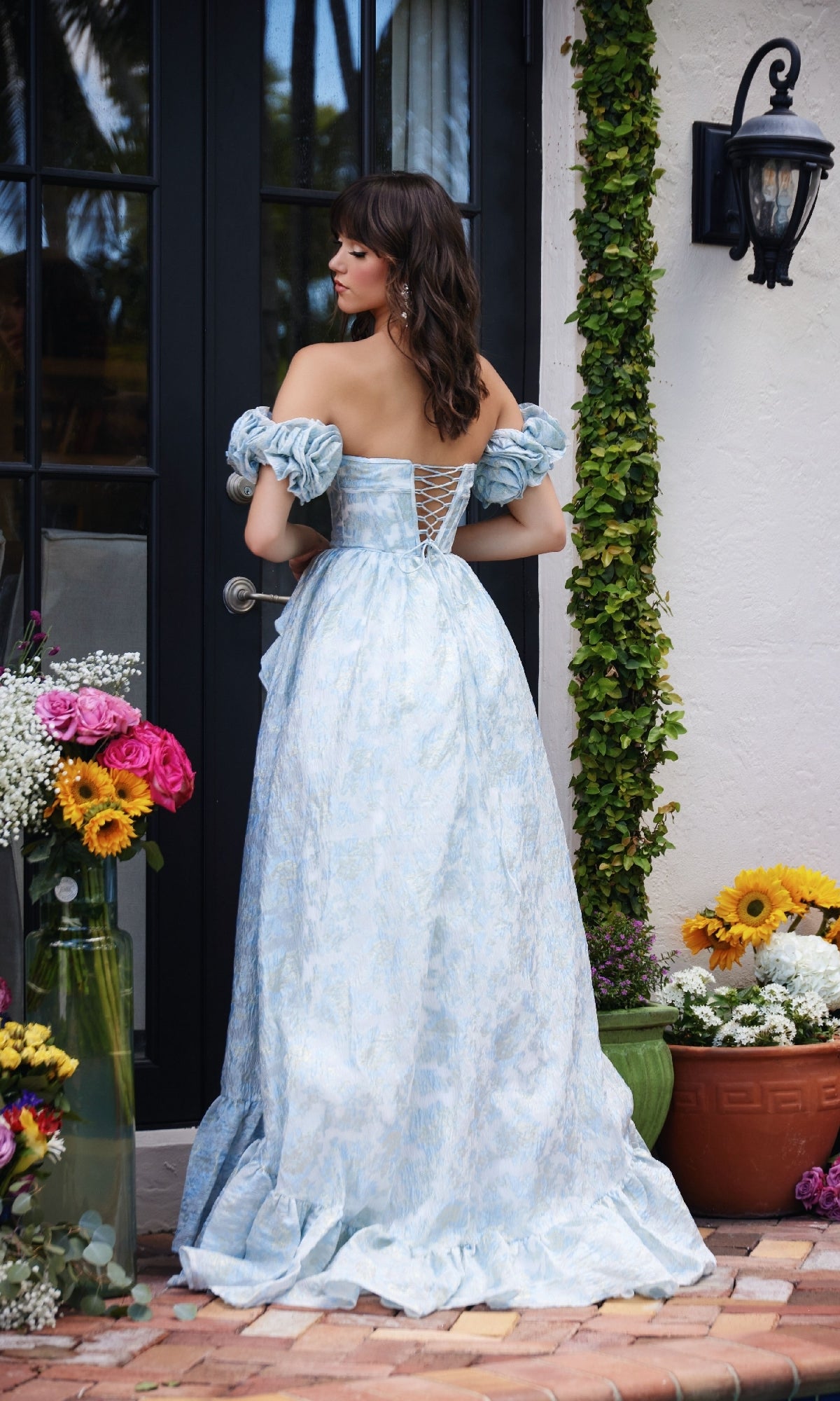 A woman stands with her back to the camera near potted flowers and a glass door, wearing the Ava Presley 40035—a light blue, off-the-shoulder, floor-length formal dress with detached puff sleeves.